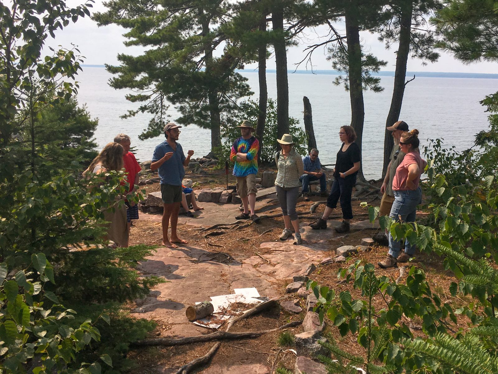 A group of visitors gathering around to listen to a resident artist talk about their practice. Rabbit Island main campfire area, sandstone ground surrounded by native white pine trees with Lake Superior in the background.